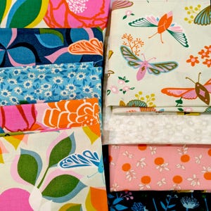 Op de afbeelding: Een verzameling gevouwen stoffen met diverse patronen. De ontwerpen bevatten kleurrijke vlinders op crème, roze met oranje cirkels, blauw met witte bloemen en grootschalige bloemenprints in roze, oranje, groen en donkerblauw.