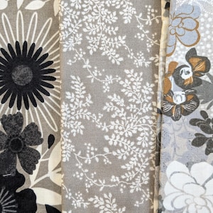 Peut inclure: Gros plan sur trois échantillons de tissu avec des motifs floraux et botaniques. Les échantillons présentent une variété de motifs, notamment de grandes fleurs noires et blanches, de délicates feuilles blanches sur fond gris et des fleurs stylisées grises, blanches et marron.
