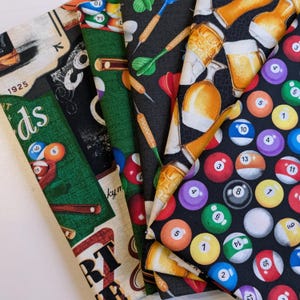 Peut inclure: Plusieurs morceaux de tissu avec des motifs sur le thème des jeux. Les motifs comprennent des boules de billard, des fléchettes, des chopes de bière et des tables de billard. Les couleurs sont le vert, le noir et le beige. Certains tissus ont du texte.