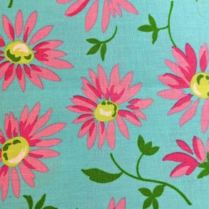May include: Close-up of a fabric with a repeating floral pattern. Large pink daisies with yellow centers are scattered across a light blue background. Green leaves and stems add to the design, creating a cheerful, summery feel.