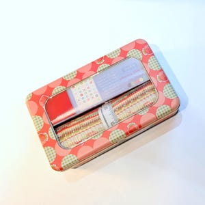 May include: A rectangular tin with a red and pink strawberry pattern holds fabric strips. The fabric strips are in a clear plastic container with a ruler printed on the side. The tin is on a white surface.