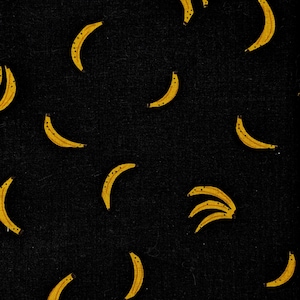 Peut inclure: Un tissu noir présentant un motif répétitif de bananes jaunes. Les bananes sont représentées dans diverses orientations, certaines comme des fruits isolés et d'autres en petits groupes, avec de petits points noirs détaillant la surface.