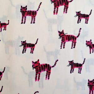 Op de afbeelding: Witte stof met een herhaald patroon van roze en zwart gestreepte kattenillustraties. De katten worden in verschillende poses afgebeeld, met snorharen en staarten. Op de achtergrond bevinden zich vage grijze katten silhouetten.