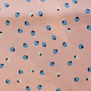 May include: Pale pink fabric featuring a repeating pattern of blue and white strawberries. Each strawberry has a black stem. The fabric has a soft texture and the strawberries are scattered across the pink background.