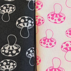 May include: Two fabric swatches featuring mushroom patterns. One is black with white mushrooms, the other is off-white with pink mushrooms. The mushroom designs have a rounded cap with polka dots and a curved stem.