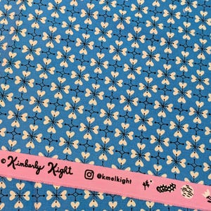 Op de afbeelding: Een helderblauwe stof met een herhalend patroon van witte vierbladige bloemvormen en dunne zwarte kruisachtige lijnen. Een roze zelfkant aan de onderkant toont de tekst Kimberly Kight, een Instagram-pictogram, @kmelkight en kleine pictogrammen.