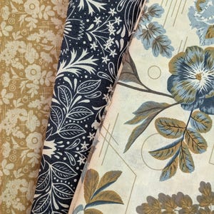 May include: Three distinct fabric swatches are displayed side by side. The left swatch features a mustard yellow floral pattern. The center swatch is dark navy blue with a white botanical print. The right swatch shows large blue and brown flowers.