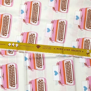 May include: A white cotton fabric featuring a repeating pattern of pink and orange vintage-style typewriters with blue hearts on the paper. A yellow selvage edge displays the text Ruby Star Society, 100% cotton, made in Japan, and darling.