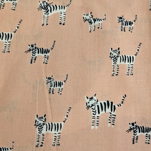 Puede incluir: Tela color melocotón con un patrón repetido de ilustraciones de tigres en blanco y negro. Los tigres están estilizados con rayas negras y bigotes. La tela es probablemente de algodón o un material similar.