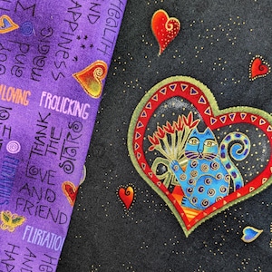 May include: Black fabric with a heart-shaped design featuring a blue cat holding flowers. The heart is outlined in red and green. Scattered hearts and stars are also present. A purple fabric with text and hearts is on the left.