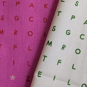 Puede incluir: Se muestran dos muestras de tela, una magenta y otra blanca. La tela magenta tiene letras rojas y estrellas blancas, mientras que la tela blanca tiene letras verdes y estrellas rosas. Ambas telas presentan un patrón repetitivo de letras.