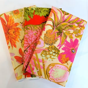 Peut inclure: Trois serviettes en tissu pliées avec des motifs floraux et fruités vibrants. Les motifs présentent de grandes fleurs, des ananas et des feuilles tropicales dans des tons d'orange, de rose, de vert et de jaune sur un fond crème. Ces serviettes conviennent à un thème tropical ou estival.