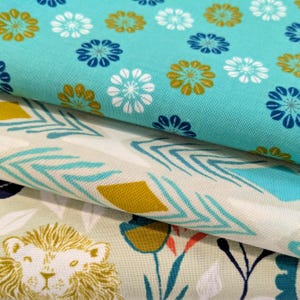 Peut inclure: Trois couches de tissu en coton empilées. La couche supérieure est turquoise avec des motifs de fleurs blanches, bleu foncé et jaune moutarde. La couche intermédiaire est crème avec des chevrons bleus et des losanges jaune moutarde. La couche inférieure est gris clair avec des visages de lion.
