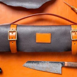 May include: A brown leather knife roll with a handle and two straps with buckles. The roll is closed and has a small orange leather patch on the front. A knife is lying on a brown surface next to the roll.