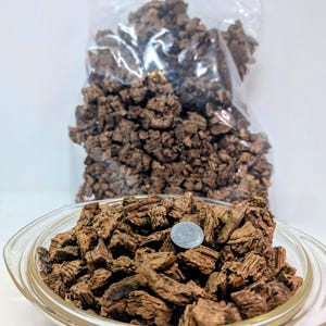 May include: A clear glass bowl filled with brown bark pieces, with a quarter coin on top. A clear plastic bag filled with similar bark pieces is in the background. The bark pieces have a rough, textured appearance.