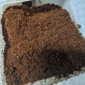 May include: A close-up of a white plastic scoop filled with brown coconut coir, a soil-like substrate made from coconut husks.