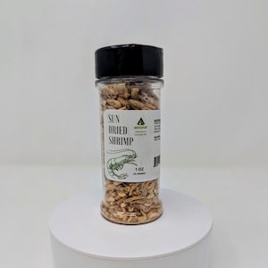 May include: A clear plastic jar filled with sun-dried shrimp, topped with a black lid. The label reads "SUN DRIED SHRIMP" with a shrimp illustration and "1 OZ (15 GRAMS)". The jar sits on a white, circular platform.