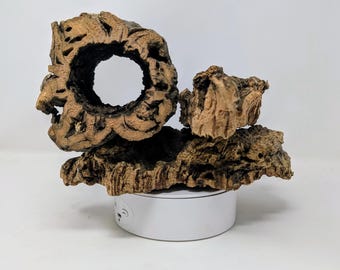 May include: A decorative piece made of natural wood bark with a large hole in the center. The bark is brown and gray with a rough texture. The piece is sitting on a white round base.