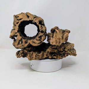 May include: A decorative piece made of natural cork bark, featuring a large hole in the center and a smaller piece of cork bark on top. The piece is mounted on a white circular base.