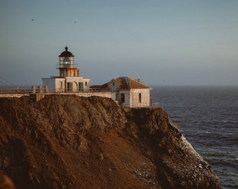 May include: A white lighthouse with a black roof sits on a cliff overlooking the ocean. The lighthouse is surrounded by a rocky shore and the water is a deep blue.