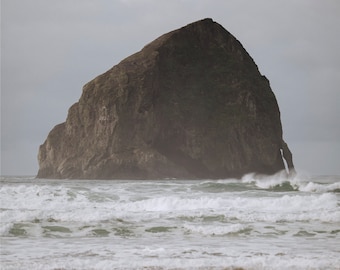 May include: A large, dark gray rock formation rises out of the ocean, with white foamy waves crashing against the shore. The sky is a muted gray, creating a dramatic and moody scene.