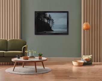 May include: A framed landscape photograph depicting a coastal scene with silhouetted trees on a cliffside. The photograph is in a black frame and hangs on a muted green wall. A round coffee table, sofa, and floor lamp are in the foreground.