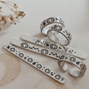 May include: Five silver rings featuring a pumpkin, leaves, and stars design. The rings are displayed on a white surface with dried flowers.