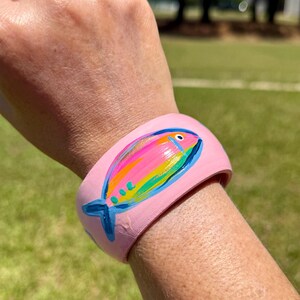 May include: A pink wooden bangle bracelet with a hand-painted fish design. The fish features a rainbow of colors, including pink, yellow, blue, and green. The bracelet is worn on a wrist.