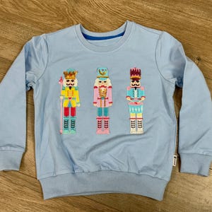 May include: A light blue sweatshirt featuring three embroidered nutcracker figures in yellow, pink, and blue. The sweatshirt has a crew neck, long sleeves, and ribbed cuffs and hem. The nutcrackers have detailed uniforms and are standing on a light blue background.