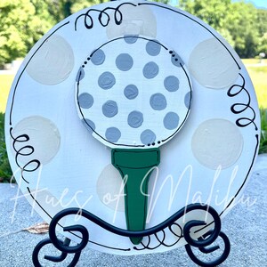May include: A white wooden round sign with a gray and white polka dot golf ball on a green tee. The sign has a black metal stand and the words "Tues of Malibu" written on it.