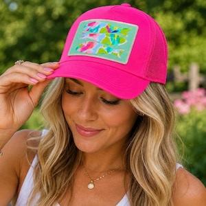 May include: A bright neon pink trucker hat featuring a rectangular patch on the front. The patch displays three stylized fish in a vibrant, multi-colored abstract pattern against a light mint green background. The hat has a mesh back panel.