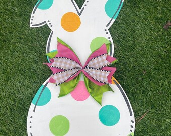 Puede incluir: Colgador de puerta de madera blanco en forma de conejo con un lazo rosa, verde y marrón. El conejo está decorado con lunares coloridos en tonos rosa, naranja, verde y azul.