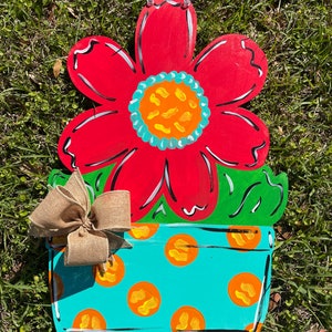 May include: A red flower with a turquoise and orange center is painted on a wooden door hanger. The flower is in a turquoise pot with orange polka dots and a brown burlap bow.