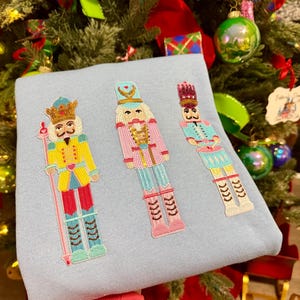 May include: A light blue sweatshirt featuring three embroidered nutcracker figures in vibrant colors. The nutcrackers have detailed uniforms with crowns, holding staffs, and standing on boots. The background includes a decorated Christmas tree.