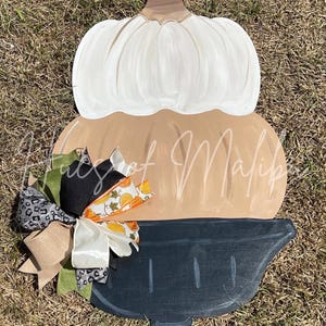 May include: A wooden pumpkin door hanger with a black pedestal base. The pumpkin is painted in shades of brown and white. A decorative bow with black, white, green, and orange ribbons is attached to the top of the pumpkin.