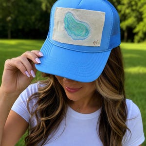 May include: A bright blue trucker hat featuring a rectangular off-white fabric patch on the front panel. The patch displays a painted illustration of a light blue and green oyster shell with the initials MK written in the bottom right corner.