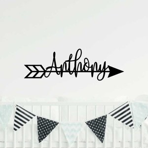 May include: Black and white arrow with the name "Anthony" written in cursive script. The arrow is on a white wall above a white crib with black and white patterned bunting.