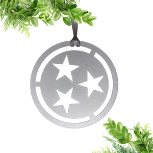 May include: A silver-colored, circular ornament featuring three white stars within a cut-out design. The ornament is suspended by a black ribbon and is decorated with green foliage. This decorative item is suitable for holiday or year-round display.