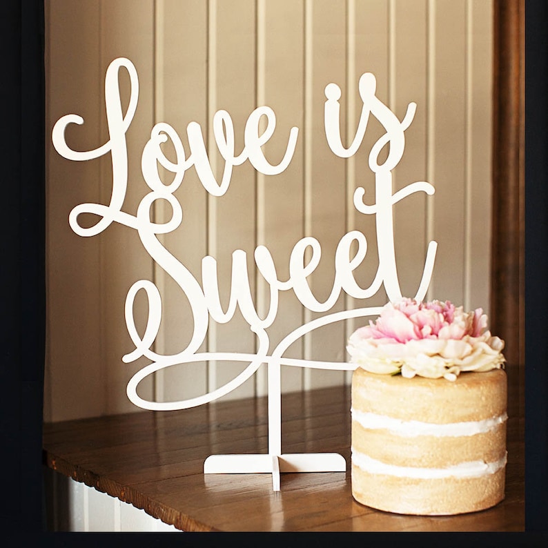 May include: White cake stand with the words "Love is Sweet" in a cursive font. The cake stand is decorated with a white cake topped with pink and white flowers.