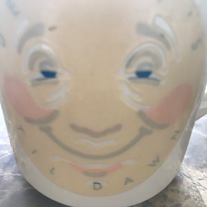 May include: A white ceramic mug featuring a printed illustration of a smiling face with rosy cheeks, grey eyebrows, and blue eyes. The text "PENNY'S" is arched above the forehead, and the letters "T I L D A W Z" are printed along the bottom edge.
