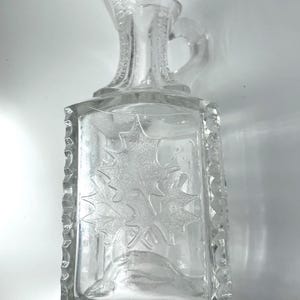May include: Clear glass pitcher with a rectangular body and a handle. The front of the pitcher features an embossed maple leaf design. The edges of the pitcher are textured. The pitcher has a spout for pouring.