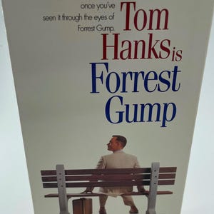 May include: A VHS tape for the movie "Forrest Gump" featuring Tom Hanks. The title is in red and blue, with the actor's name prominently displayed. The image shows a man sitting on a bench with a suitcase, evoking a sense of nostalgia.