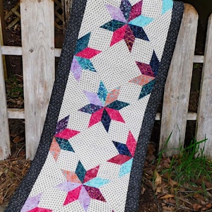 Może przedstawiać: Biały bieżnik stołowy z czarną obwódką i powtarzającym się wzorem kolorowych gwiazd. Gwiazdy są wykonane z różnych tkanin i mają patchworkowy wzór.