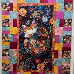 May include: A rectangular quilt featuring a central panel of a sleeping cat surrounded by colorful flowers on a black background. The border consists of a patchwork grid of squares in shades of pink, yellow, and blue, interspersed with cat-themed fabric.