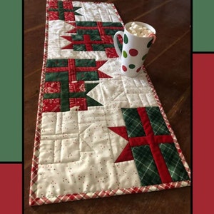 May include: A quilted table runner with a red and green plaid pattern and a white background with red polka dots. The runner features a design of wrapped presents with red bows. The text "Gift Wrapped" is at the top of the image. The text "Molly Cook for Villa Rosa Designs" is at the bottom of the image.