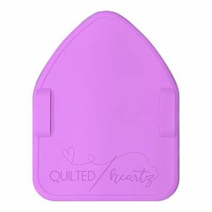Könnte beinhalten: Eine lavendelfarbene Nähmatte mit hausförmiger Oberseite. Die Matte hat zwei kleine, rechteckige Laschen an den Seiten und die Worte "QUILTED heartz" mit einem Herz und einem Stichdesign. Die Matte ist für Quilt- und Nähprojekte konzipiert.