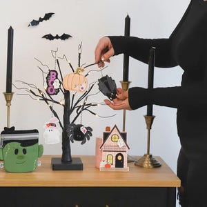 May include: Halloween-themed decorations on a wooden table. A black tree with hanging ornaments, including a pumpkin, ghost, and spider. Black candles in gold holders and a decorative house are also present. A person is holding a bat ornament.
