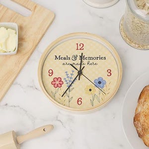 May include: A round, light-colored clock with the text "Meals & Memories are made here." The clock face features floral designs and the numbers 12, 3, 6, and 9 in red. The clock is surrounded by a light wooden frame.