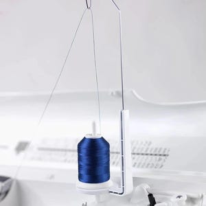 May include: A spool of vibrant blue thread is suspended by a white and silver thread guide, ready for sewing. The thread is tightly wound on a white spool, and the guide is attached to a white machine. The scene is set against a soft, neutral background.