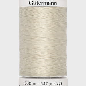 Könnte beinhalten: Eine Spule cremefarbenes Gütermann-Garn. Die Spule hat einen weißen Deckel mit dem Markennamen "Gütermann" in Schwarz aufgedruckt. Auf der Basis der Spule steht ebenfalls in Weiß "500 m - 547 yds/vgs". Das Garn ist eng gewickelt.